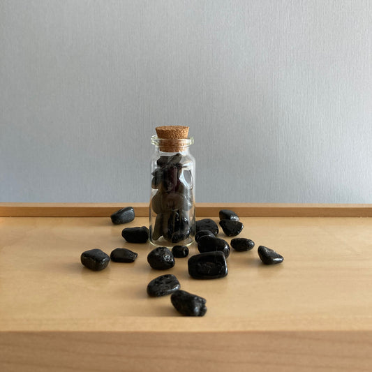 Bottle of Black Tourmaline chips sitting on a wooden box surrounded by loose Black Tourmaline chips